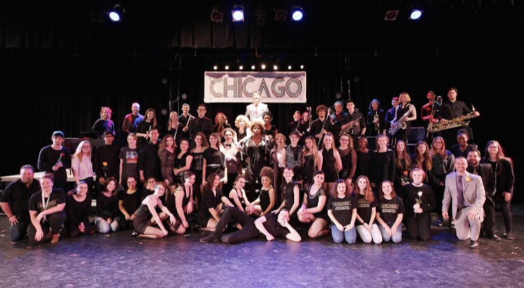 Chicago cast poses in costume on set with crew and orchestra.
